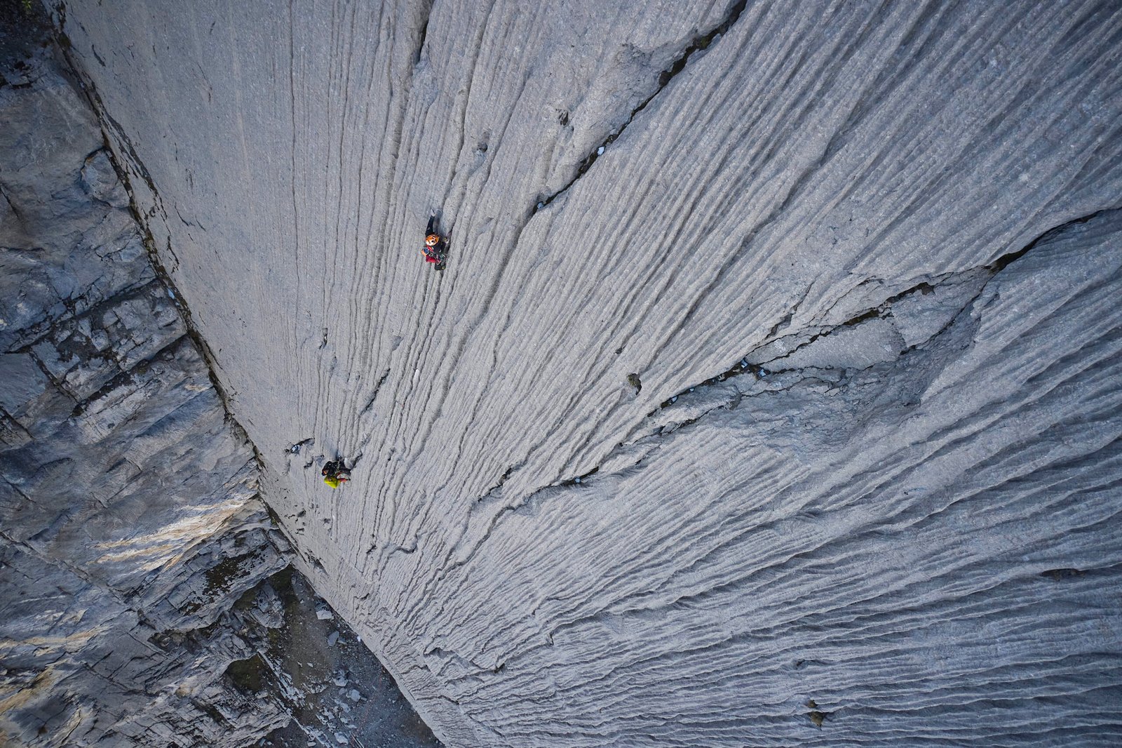 Two roped climbers tiny against a vast striated big-wall, shot from above. By Alexander Estrada.