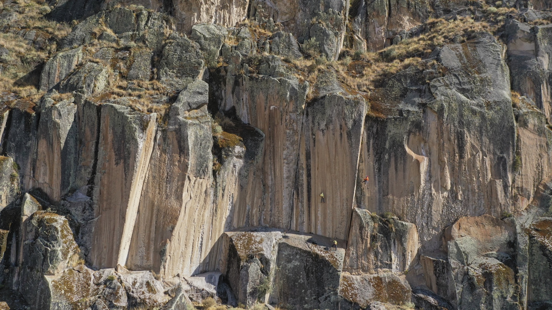 Two tiny climbers scale a vast tan and gray volcanic rock face streaked with vertical fissures in Peru's Rurec valley.