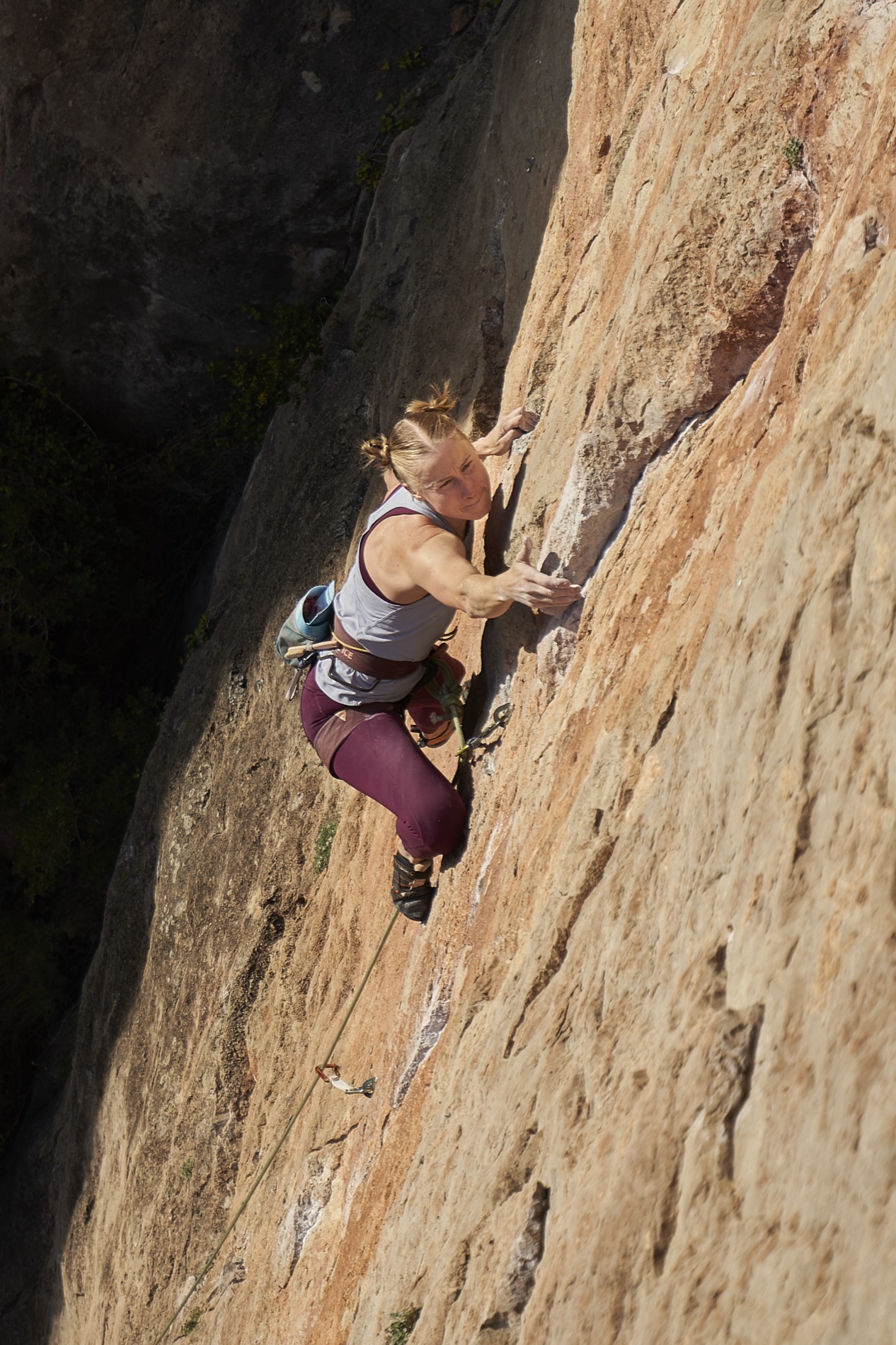 A blonde woman in a white tank top and maroon tights crimps on sunlit orange limestone, reaching high on a steep Siurana wall.