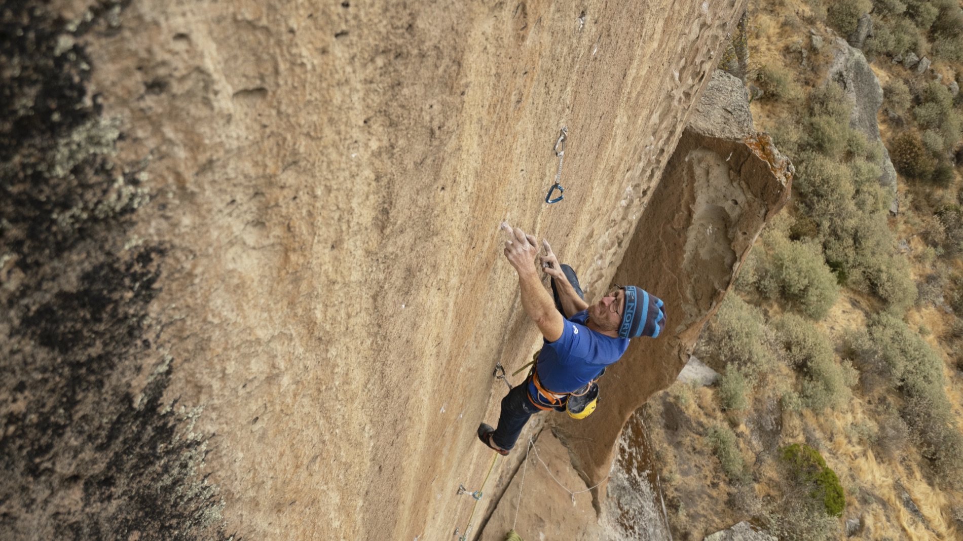Iker Pou in a blue shirt and beanie reaches for a crimp on a sheer sunlit granite face, dry Peruvian hillside below. Shot by Alexander Estrada.