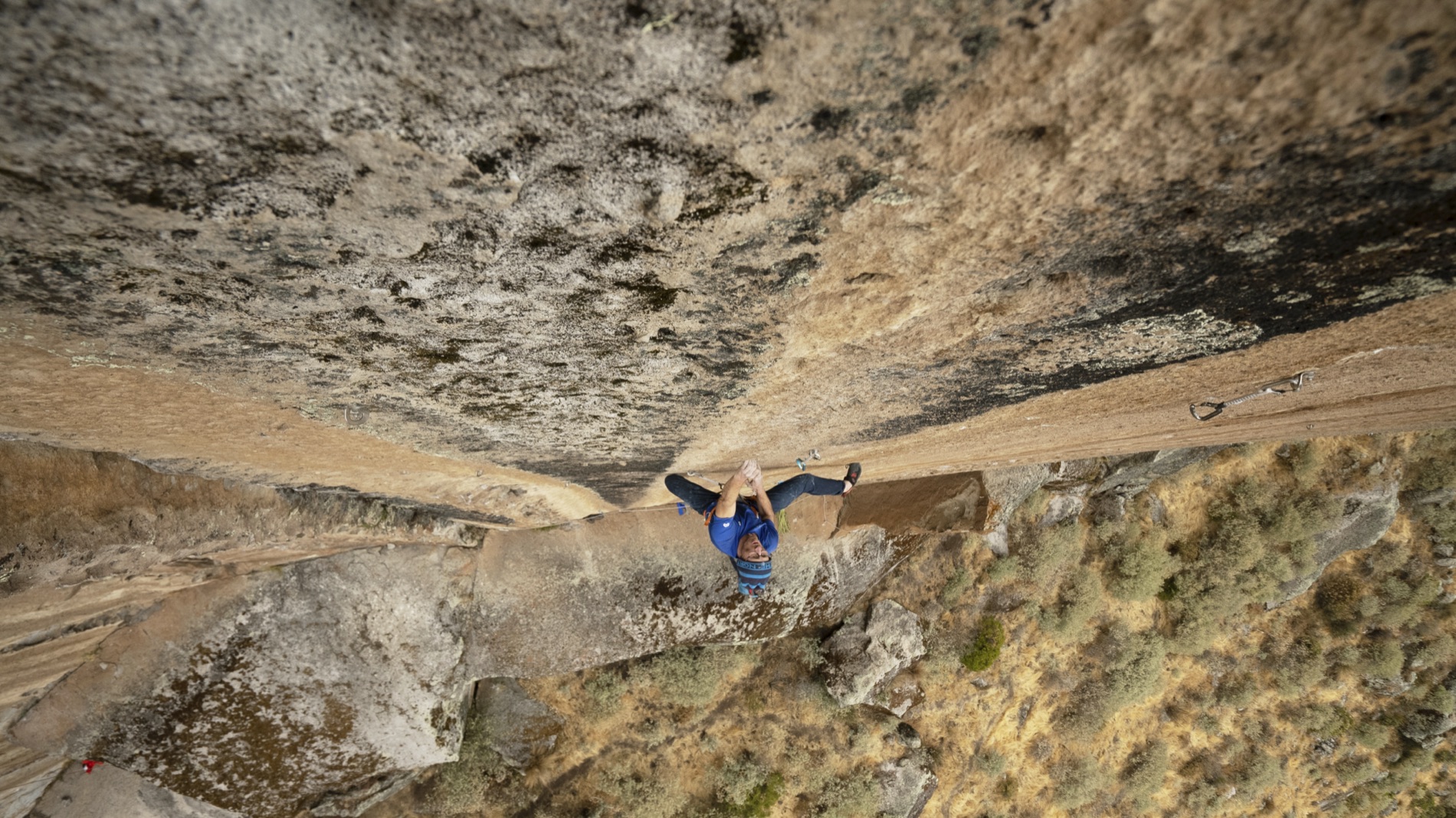Overhead view of Iker Pou in blue splayed across a steep granite slab, feet stemmed wide, looking up toward the camera.