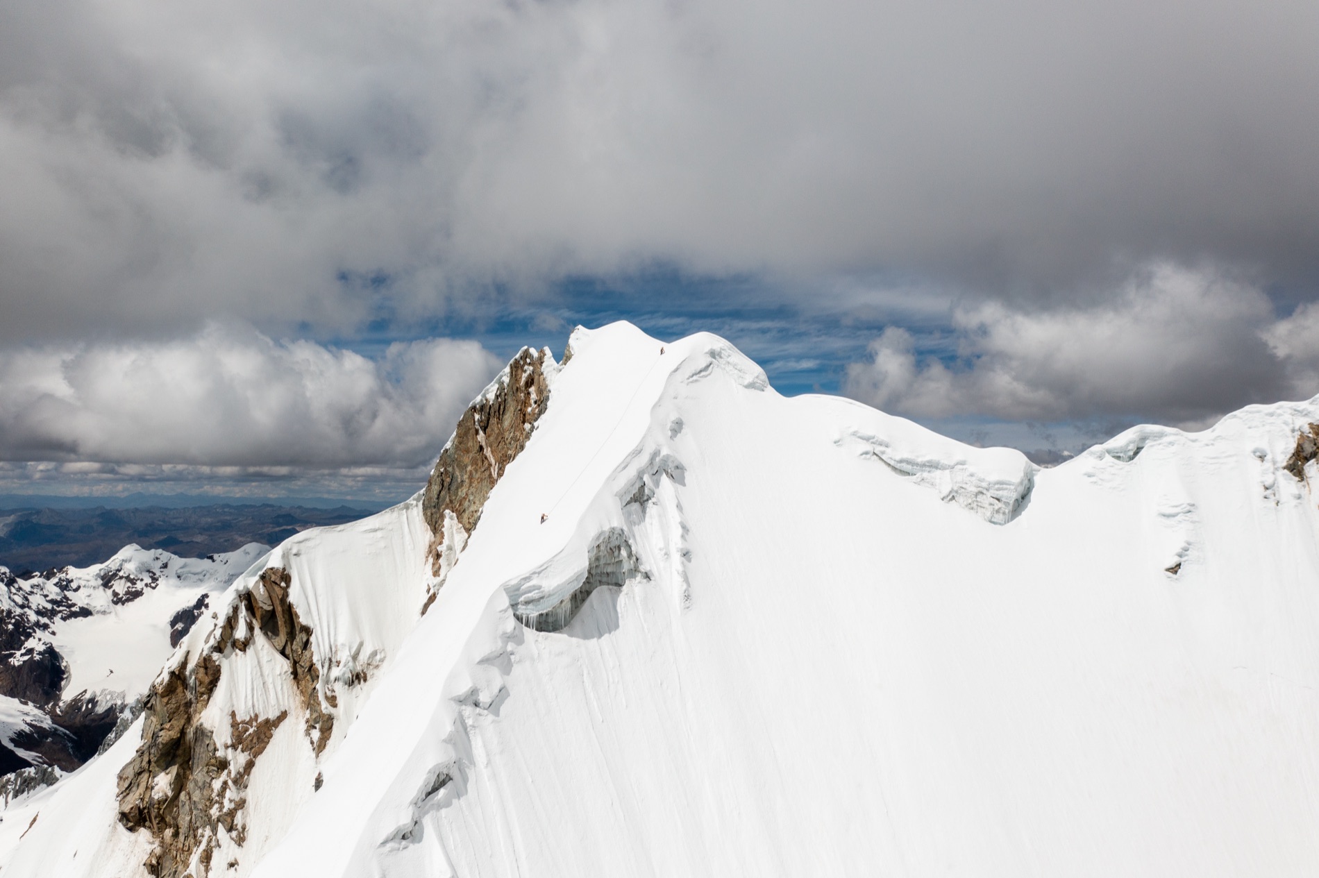 A snow-plastered Andean summit ridge with a tiny climber on the corniced crest under heavy gray clouds in the Cordillera Blanca.