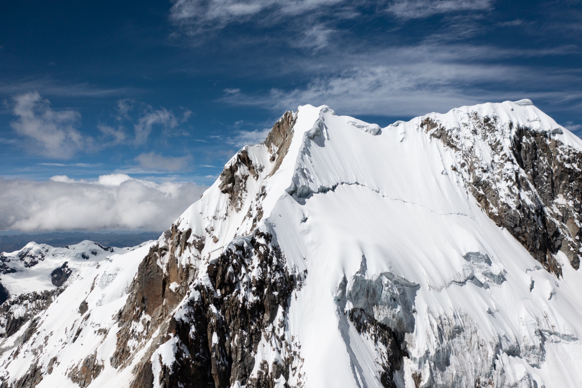 A sharp snow and rock summit of the Cordillera Blanca rises against deep blue sky with wispy clouds. Shot by Alexander Estrada.