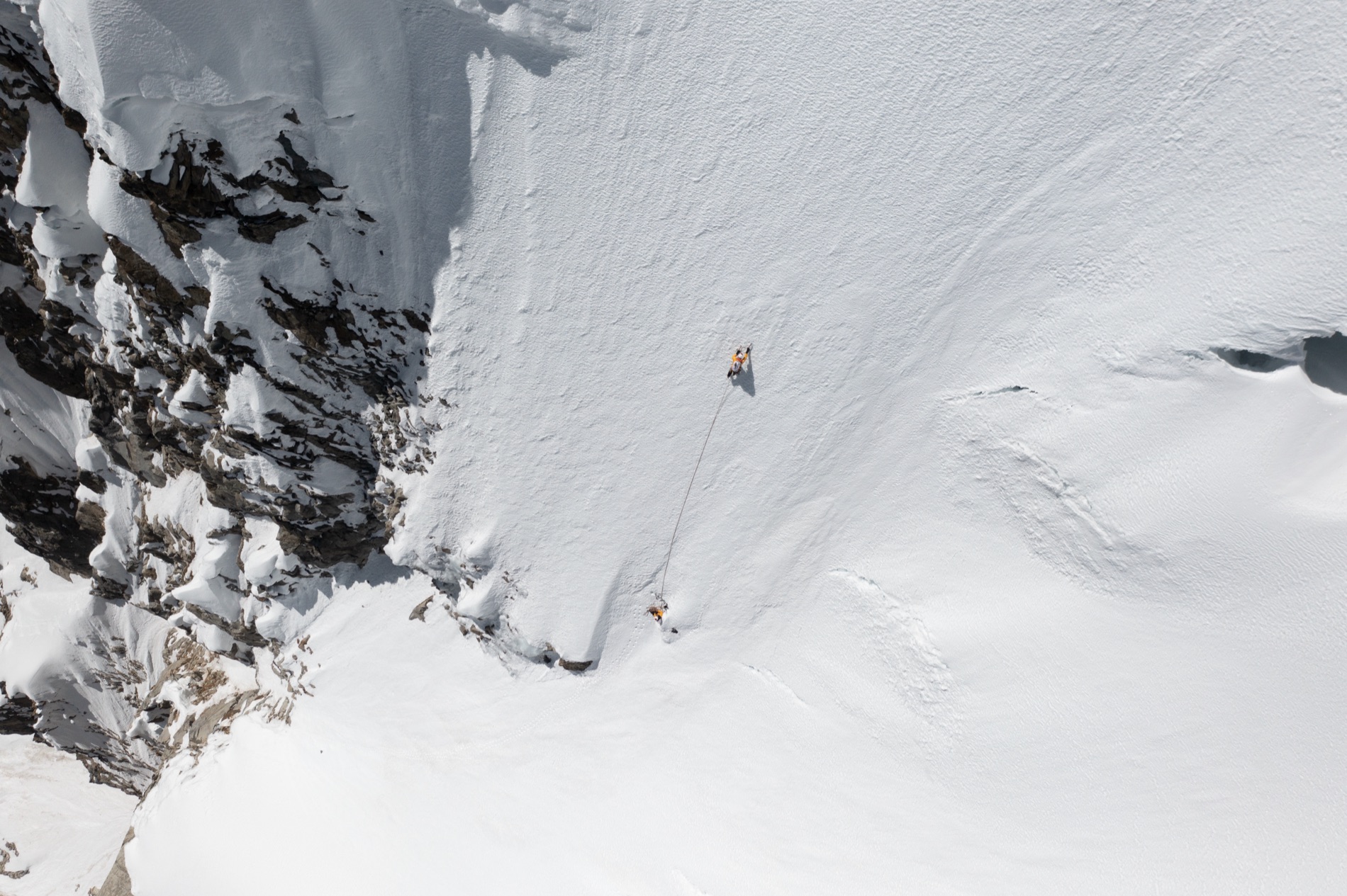 Aerial view of two roped climbers in orange crossing a vast white snow slope beside dark rock in the Cordillera Blanca.