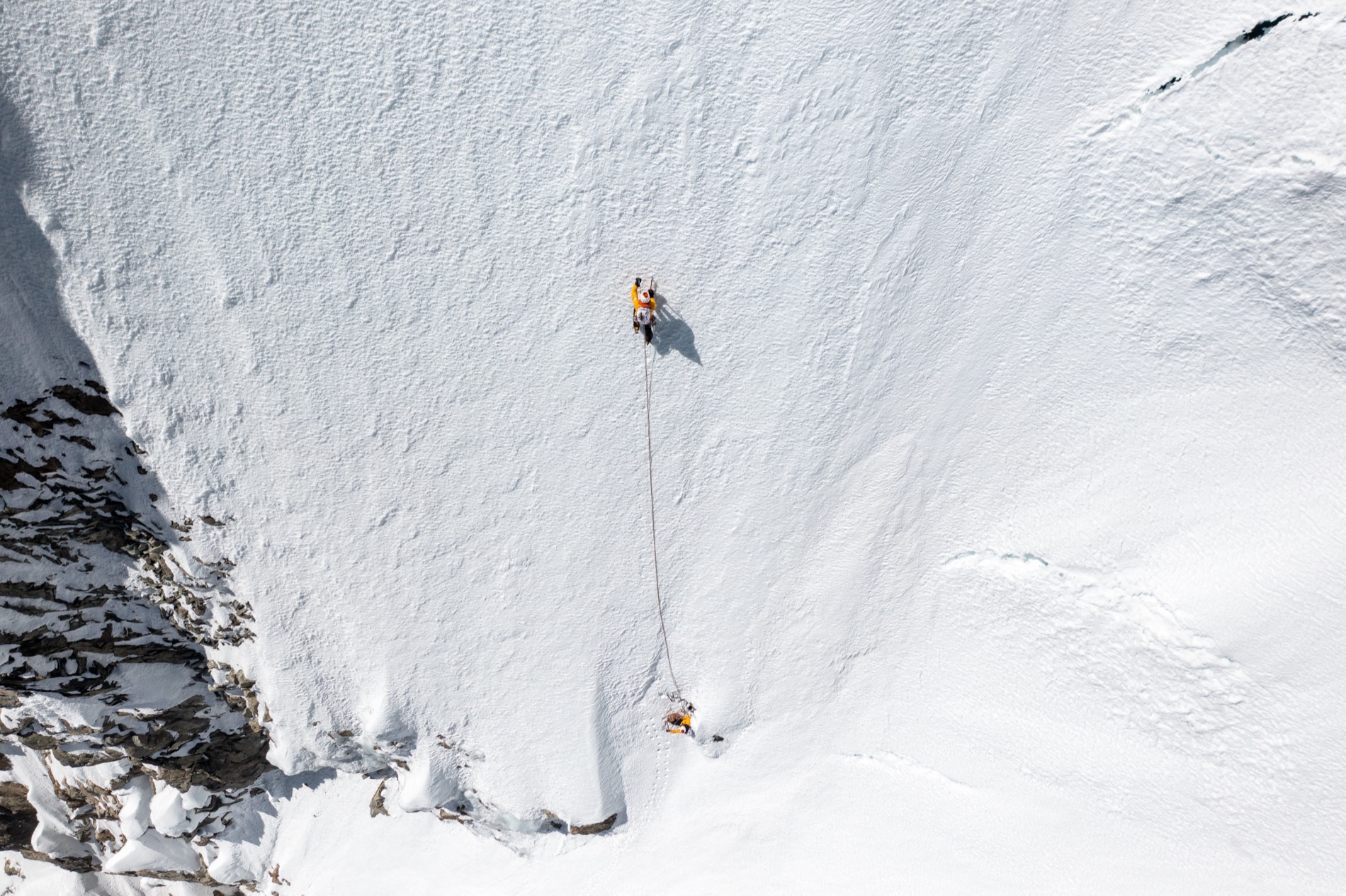 Two roped mountaineers, one in orange, climb a steep sunlit snowfield high on an Andean peak. By Alexander Estrada.