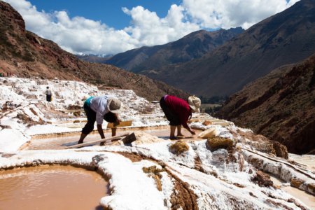 Two workers harvest salt by hand on white terraced ponds in a sunlit mountain valley