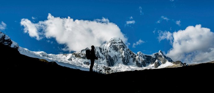 Lone hiker silhouetted against jagged snowcapped peaks and brilliant blue sky; by Alexander Estrada