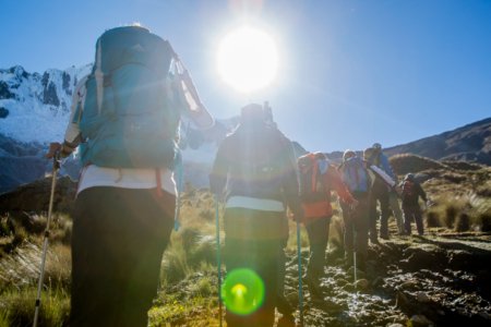 Backpackers hike a sunlit alpine trail into bright sun flare with mountains ahead