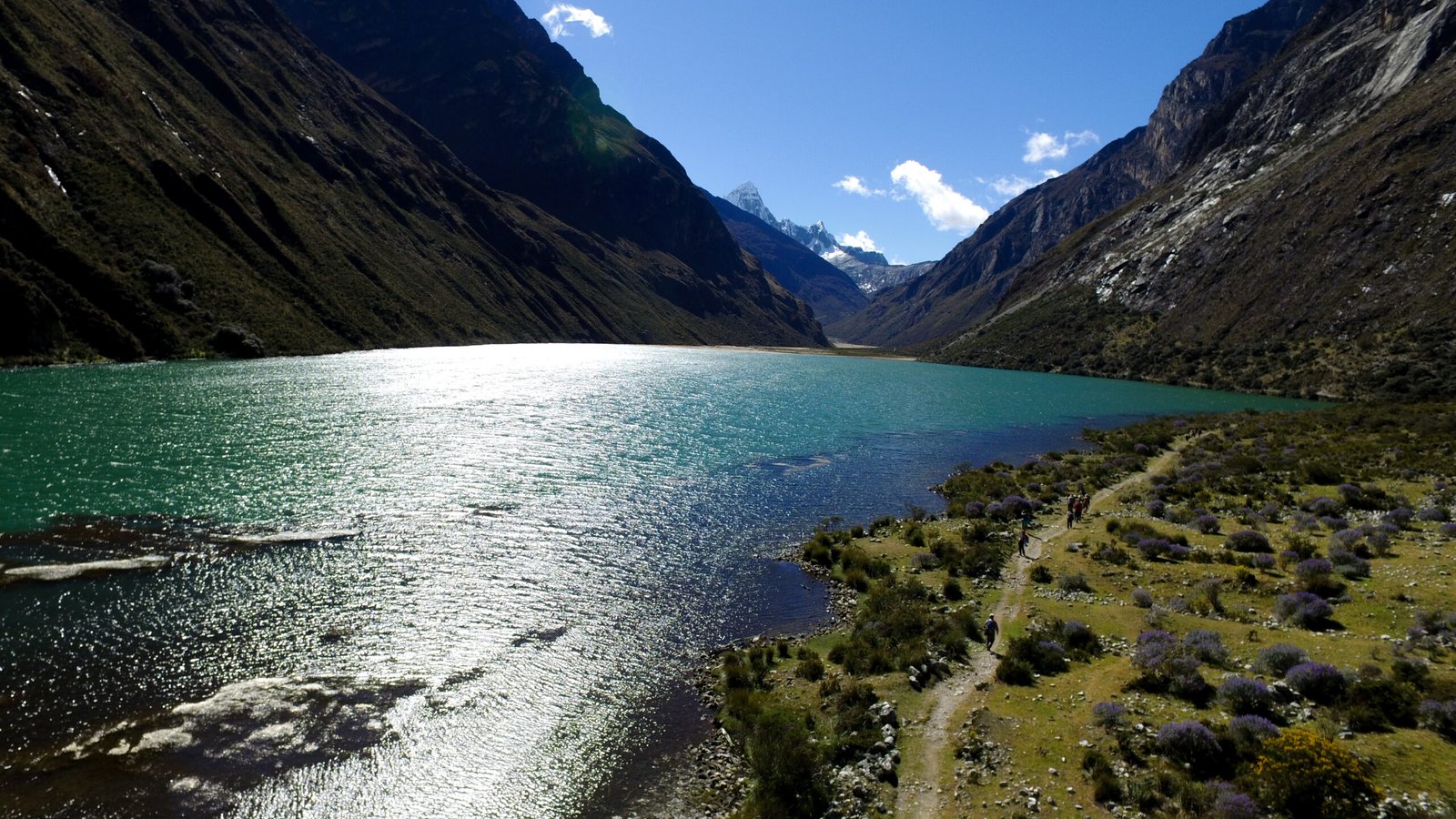 Drone view of a brilliant turquoise glacial lake and trail beneath towering valley walls; by Alexander Estrada