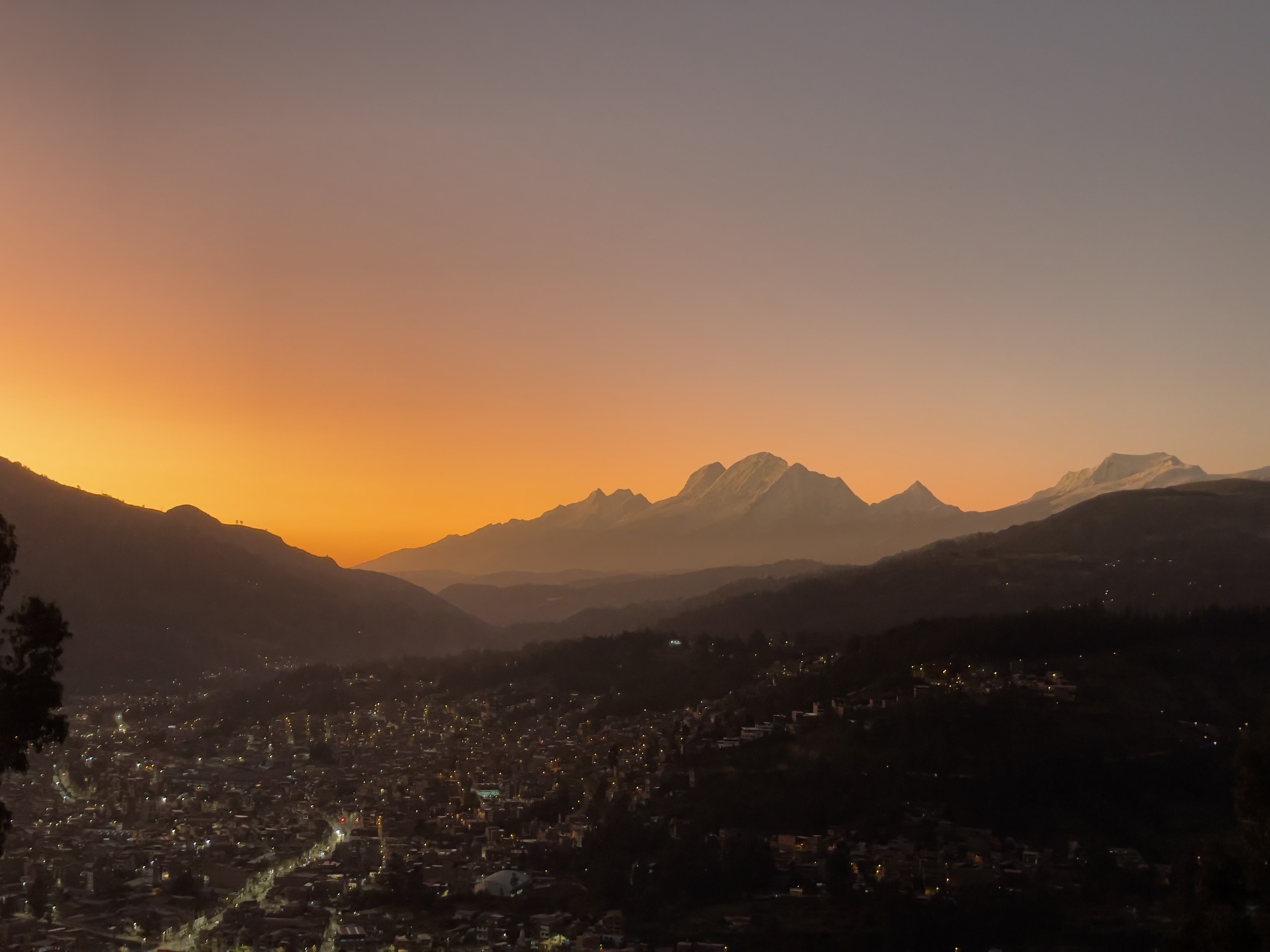 Snow-capped Andean peaks glow at dusk above a valley city dotted with lights under an orange sunset sky.