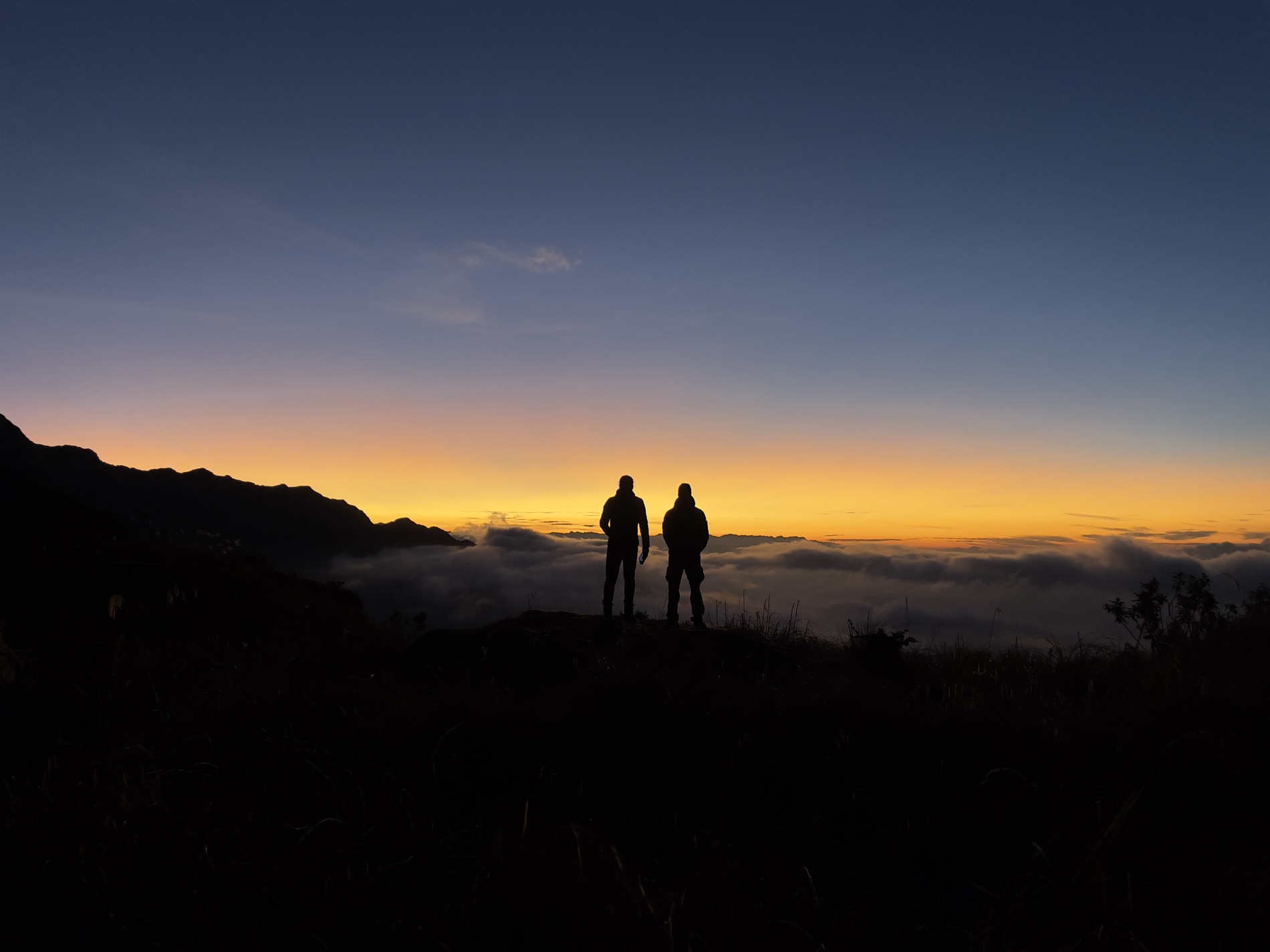 Two hikers stand in silhouette on a ridge above a sea of clouds at sunrise. By Alexander Estrada.