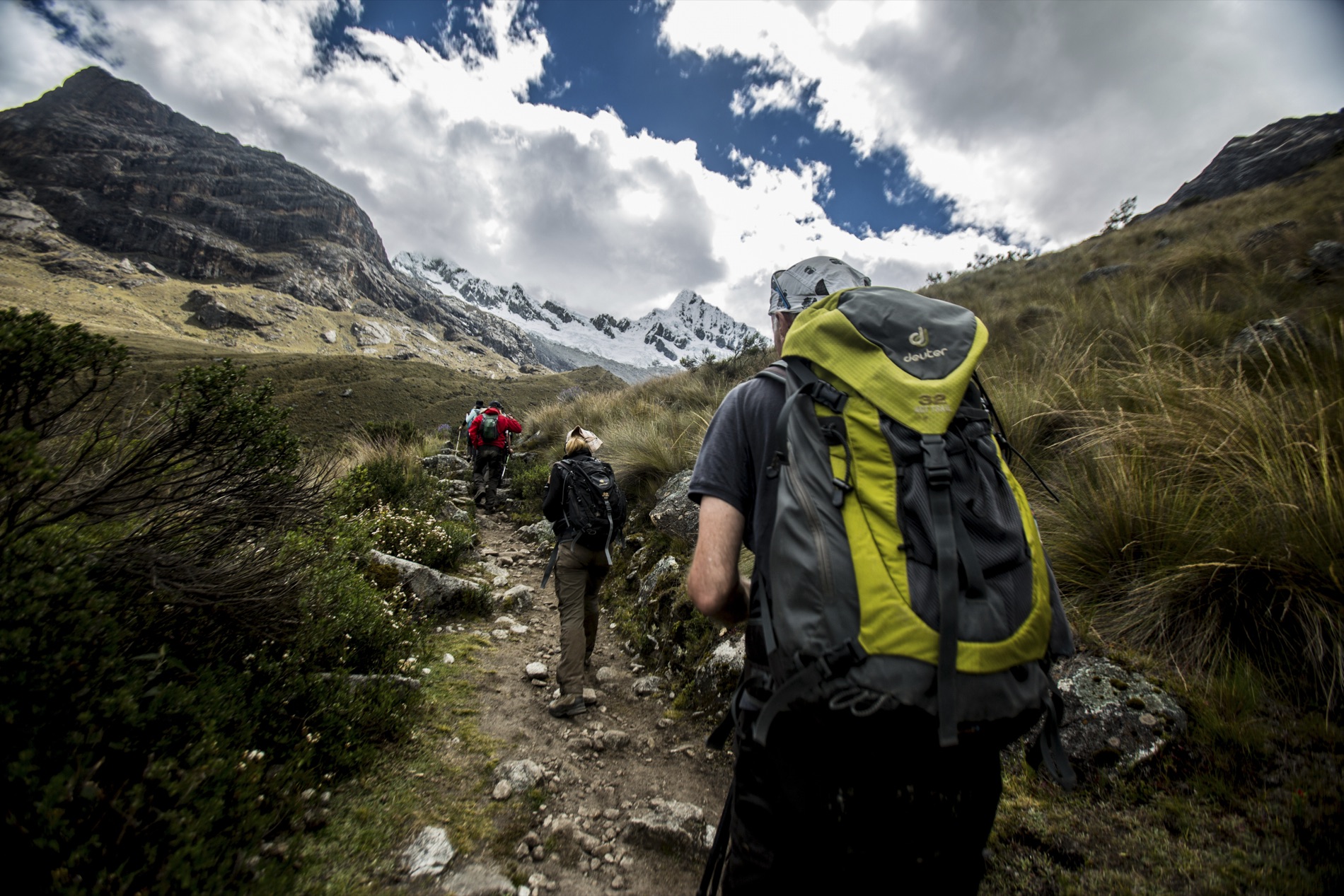 Trekkers with backpacks climb a rocky trail toward snow-capped Andean peaks under dramatic white clouds. Shot by Alexander Estrada.