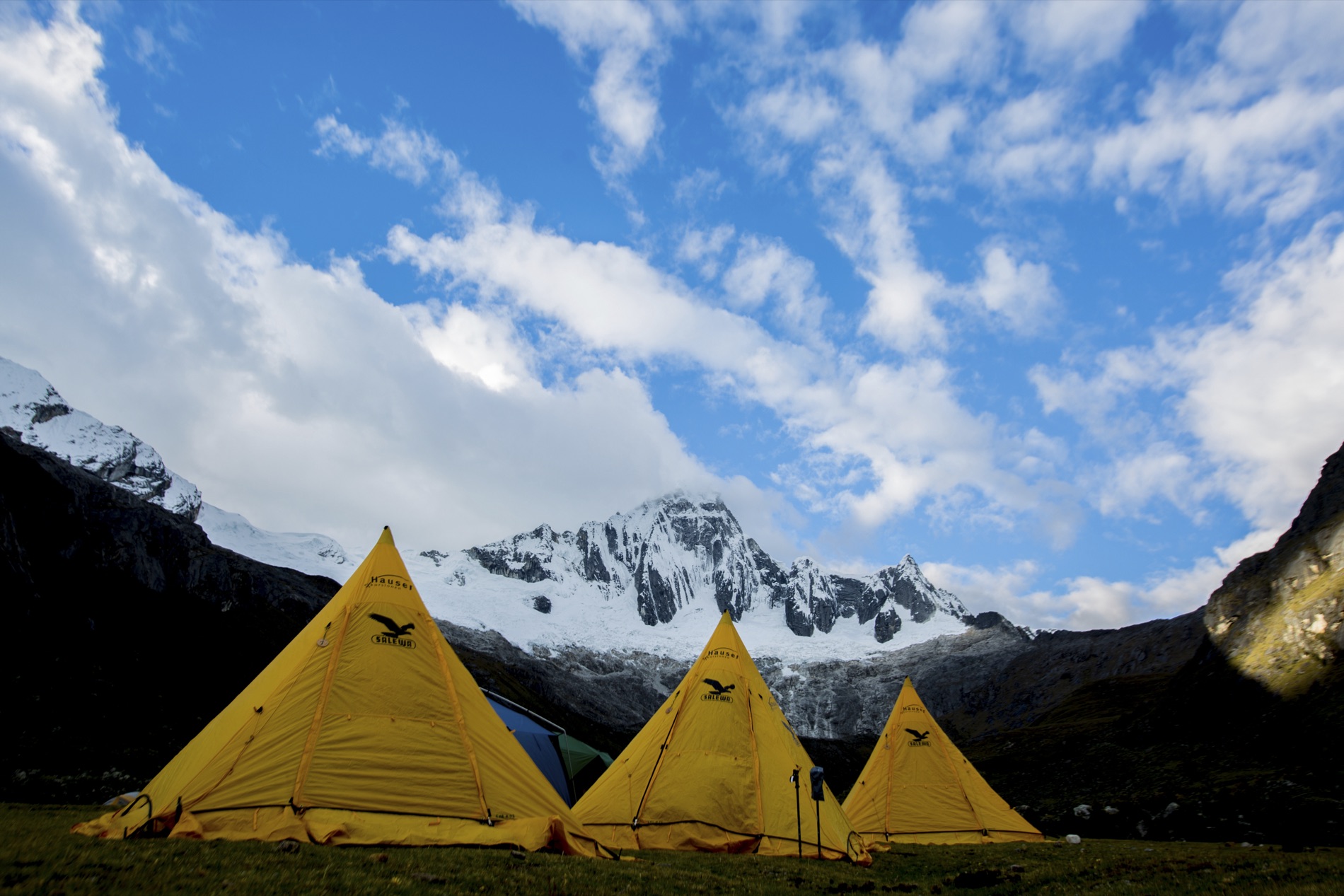 Three yellow pyramid tents stand on a grassy meadow below a snow-covered peak and blue sky. By Alexander Estrada.