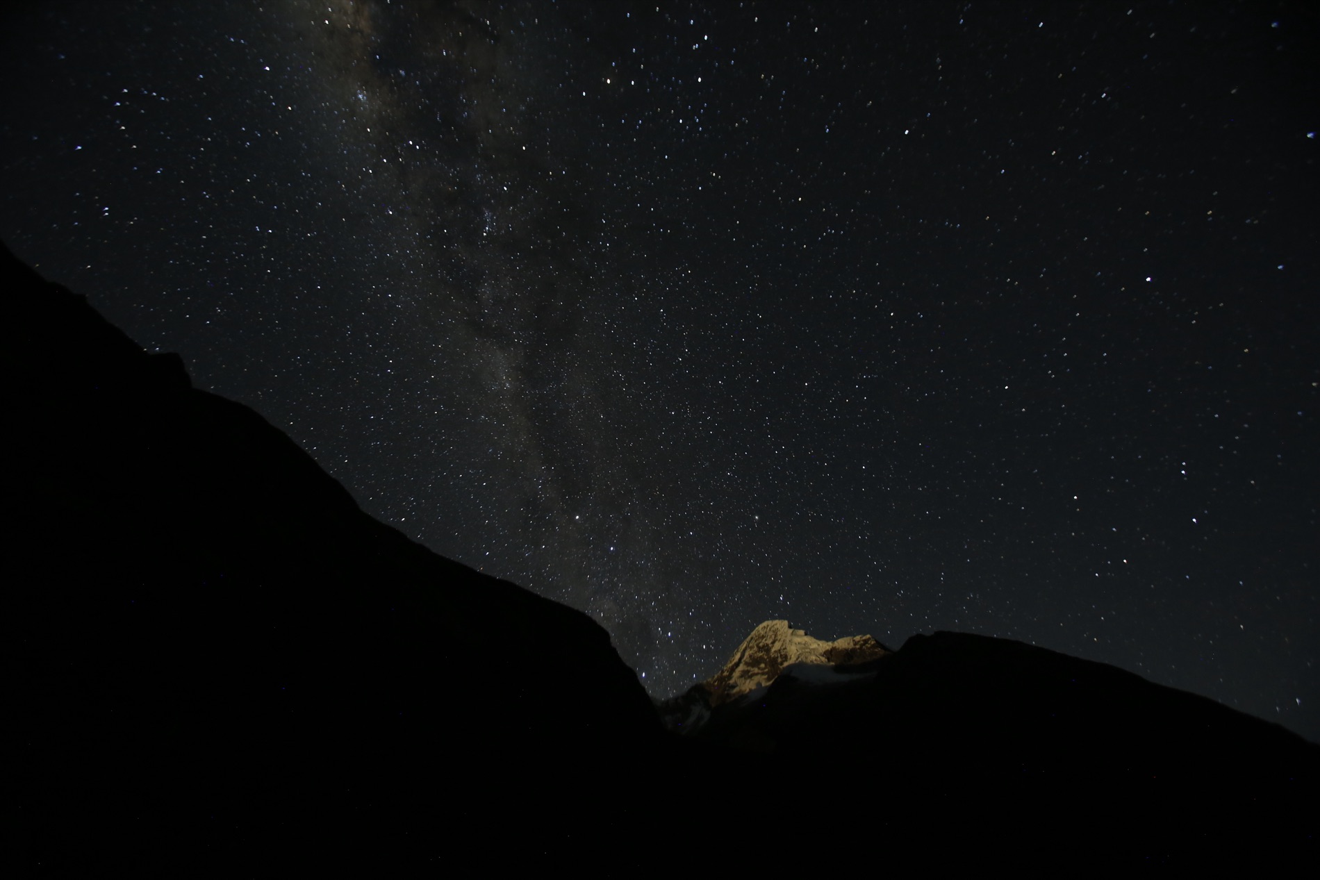 The Milky Way arcs across a star-filled night sky above a faintly lit snow peak framed by dark ridges.