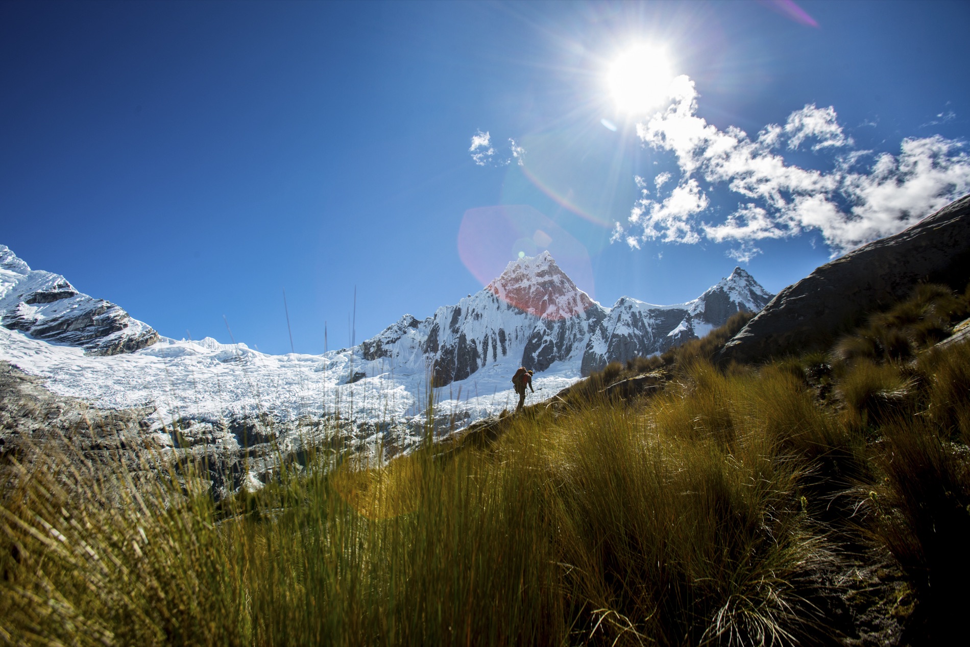 A lone hiker crosses a golden grassy slope beneath jagged snow peaks and a glacier as the sun flares overhead.
