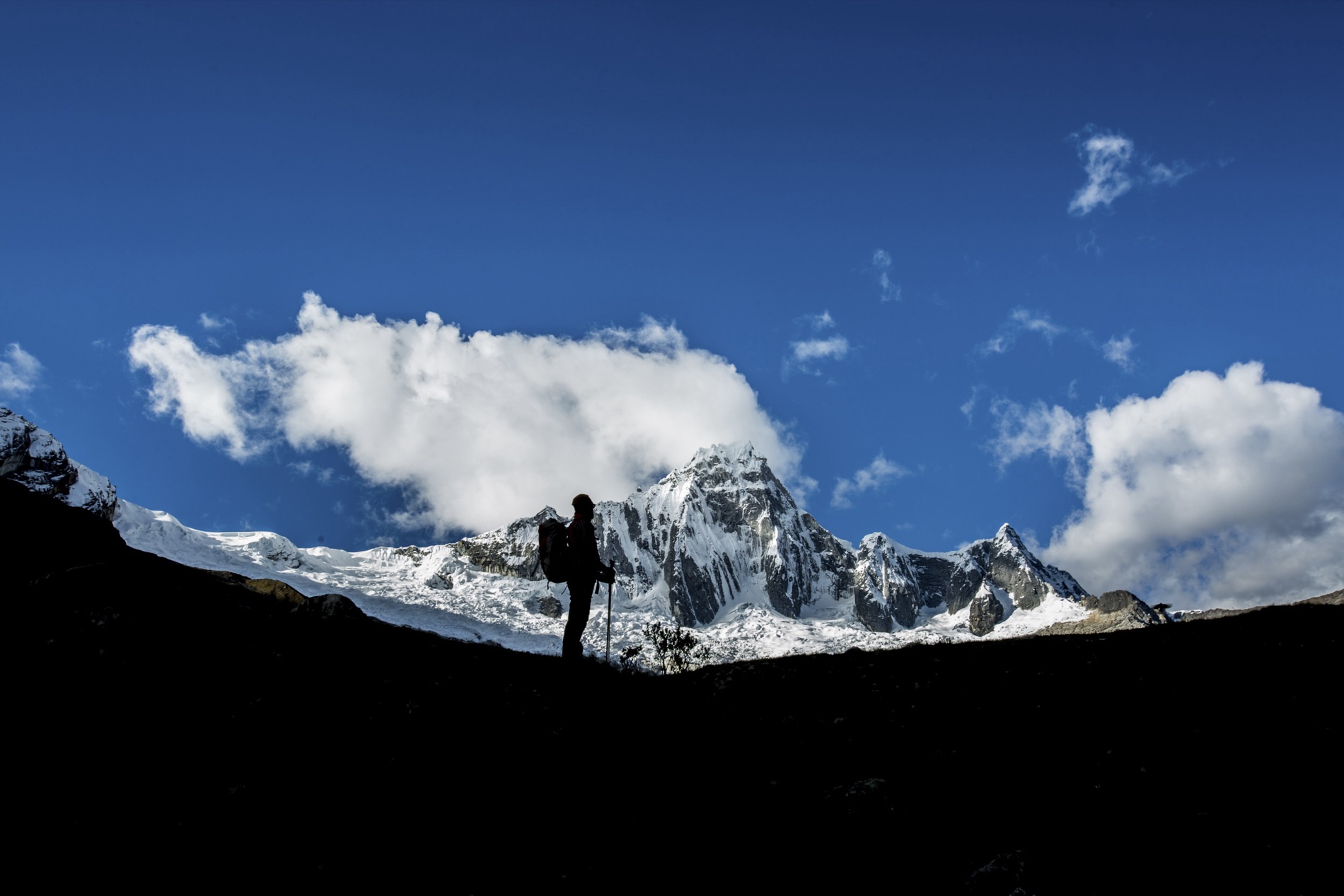 A backpacker stands in silhouette on a dark ridge facing a snow-capped Andean peak under blue sky and clouds. Shot by Alexander Estrada.