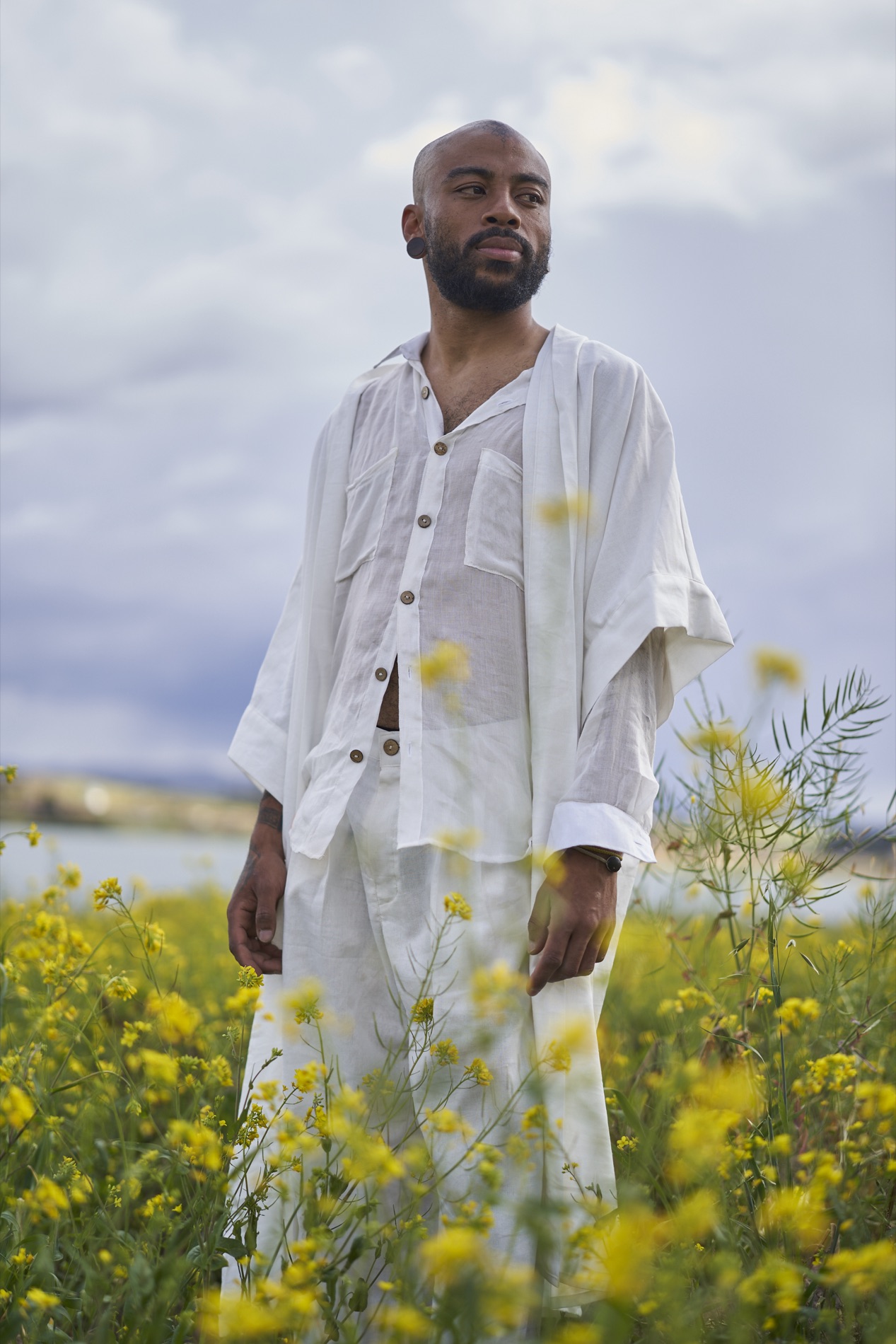 Environmental portrait of a bearded man in a loose white linen shirt and trousers standing in a field of yellow flowers under a cloudy sky. Portrait by Alexander Estrada.