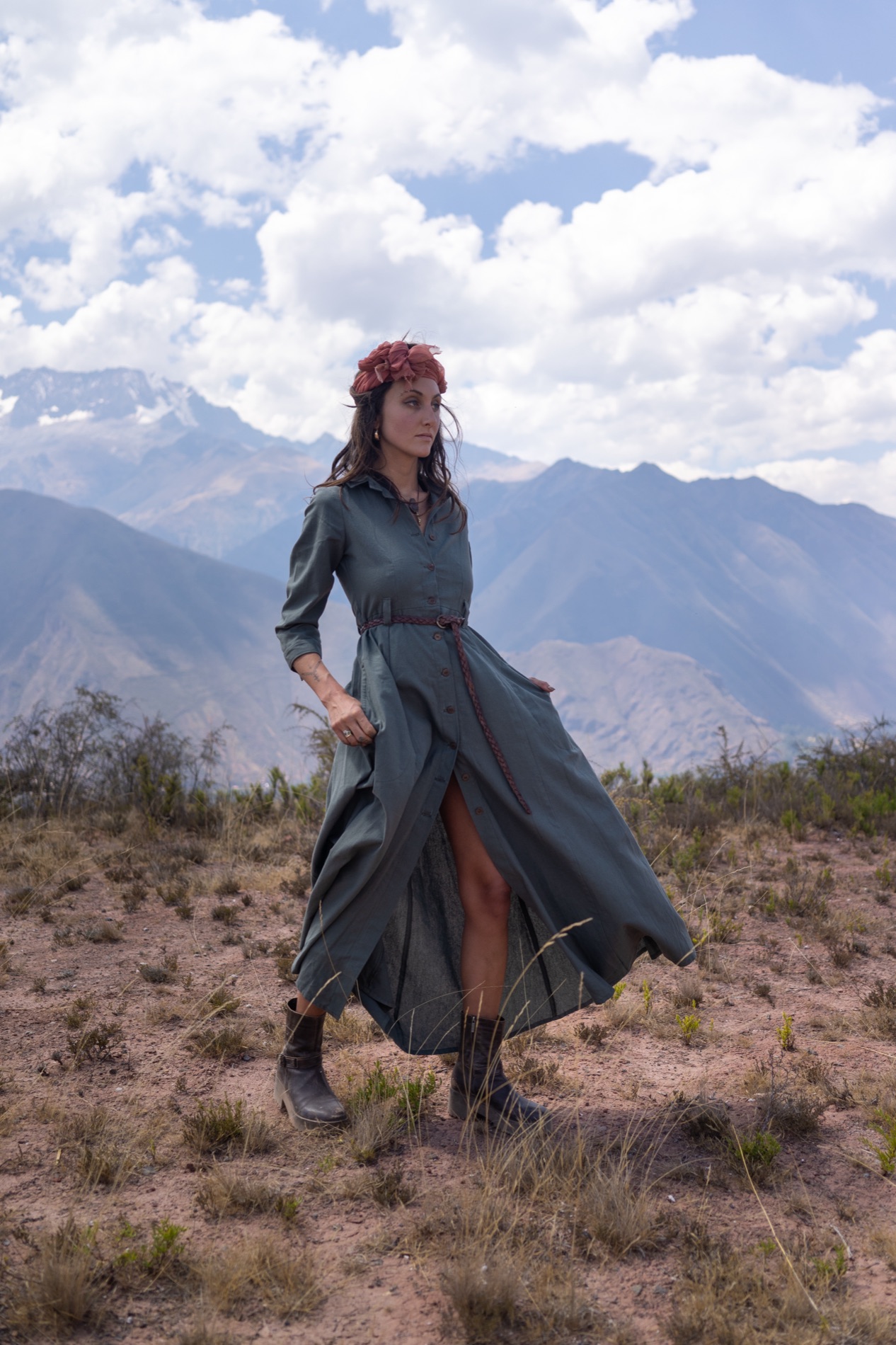 Outdoor portrait of a woman in a long gray-green dress and boots walking across a dusty mountain plateau with peaks and clouds behind her.