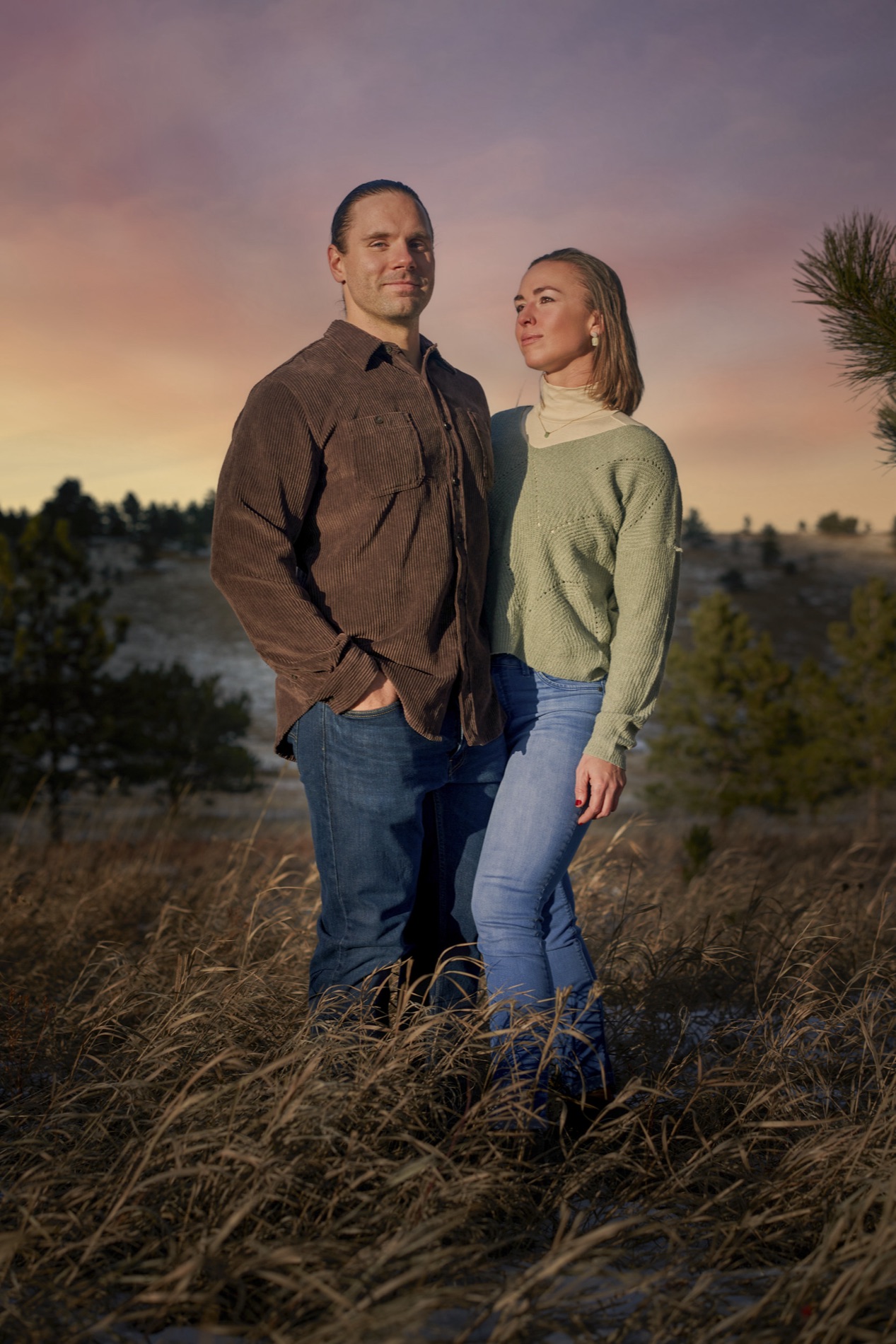 Environmental portrait of a couple standing in tall golden grass at dusk, the man in a brown corduroy shirt and the woman in a sage sweater. Portrait by Alexander Estrada.