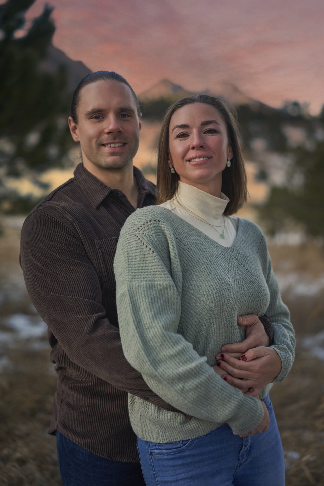 Outdoor portrait of a smiling couple embracing in a mountain meadow at sunset, the woman in a sage sweater and the man in a brown corduroy shirt.