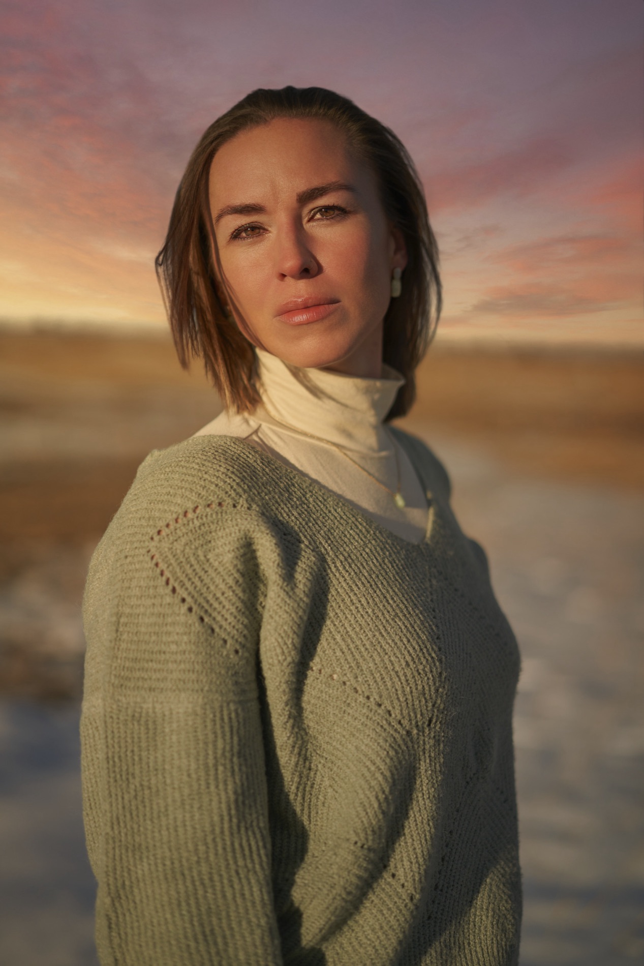 Golden-hour portrait of a young woman in a sage sweater and cream turtleneck standing in an open field under a pink sunset sky. Portrait by Alexander Estrada.