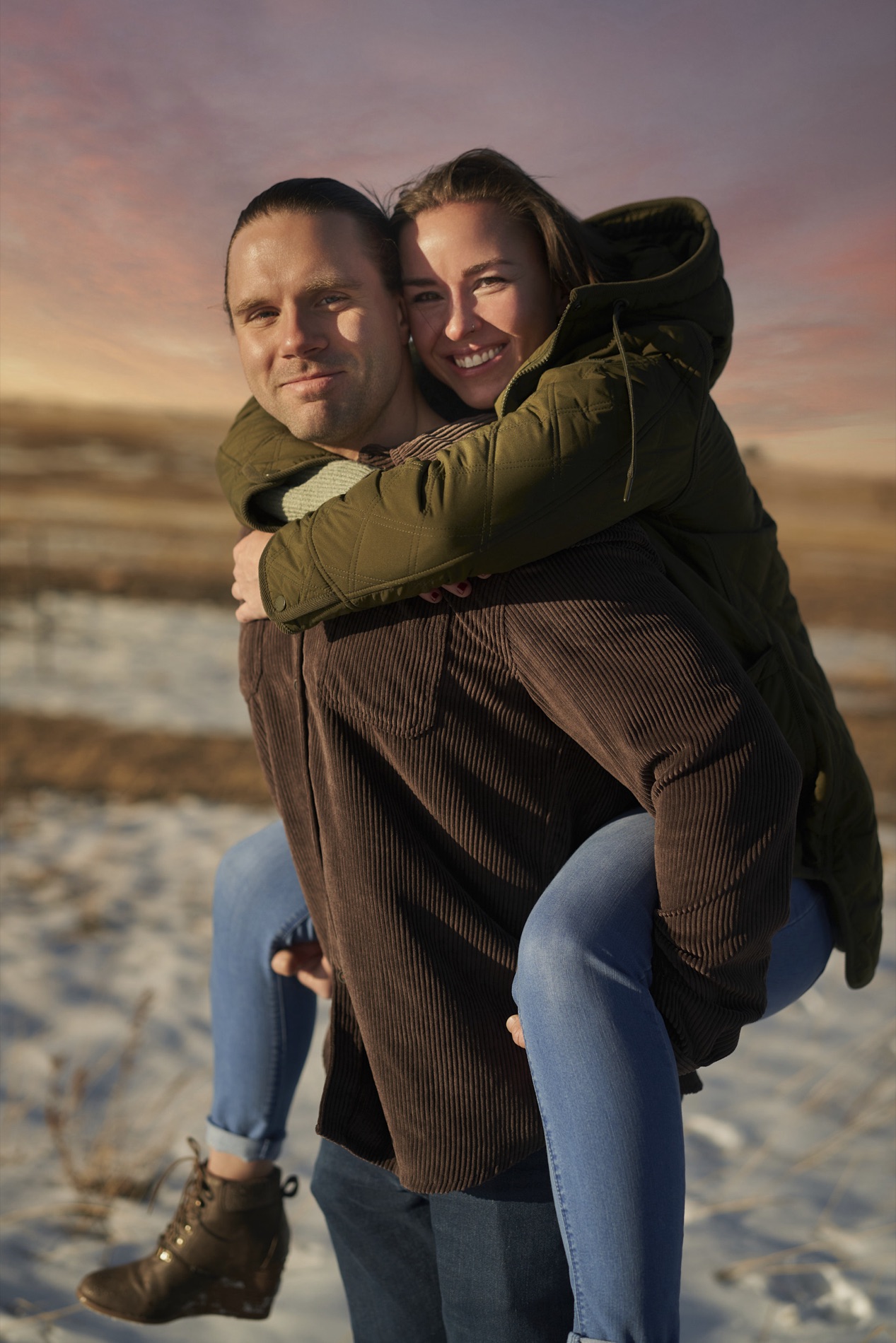 Documentary portrait of a smiling couple giving a piggyback ride across a snowy field at sunset, the woman in a green jacket and the man in a brown shirt.