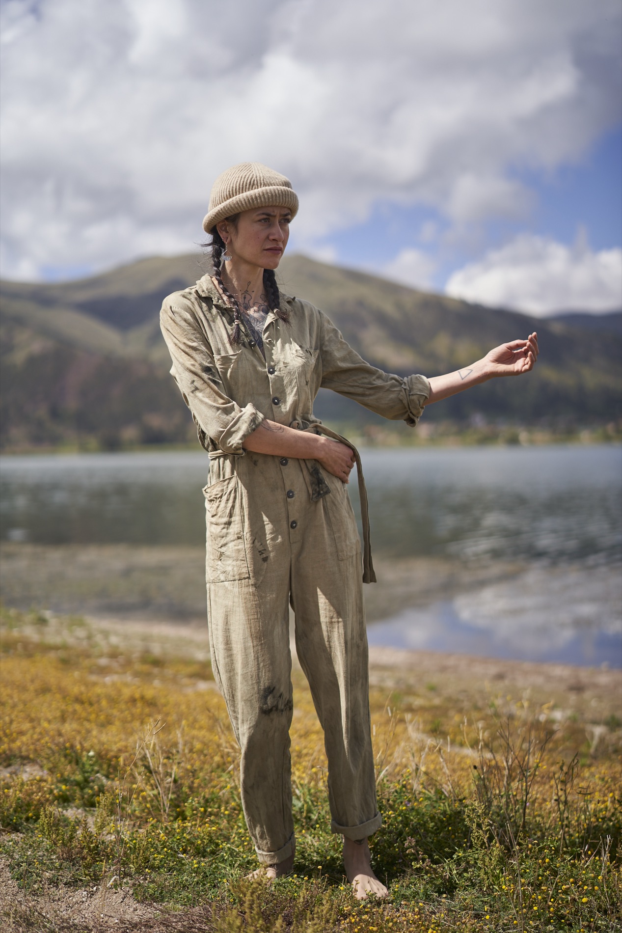 Environmental portrait of a woman in a dusty tan coverall and knit cap standing barefoot on grassy ground beside a calm lake and green mountains.