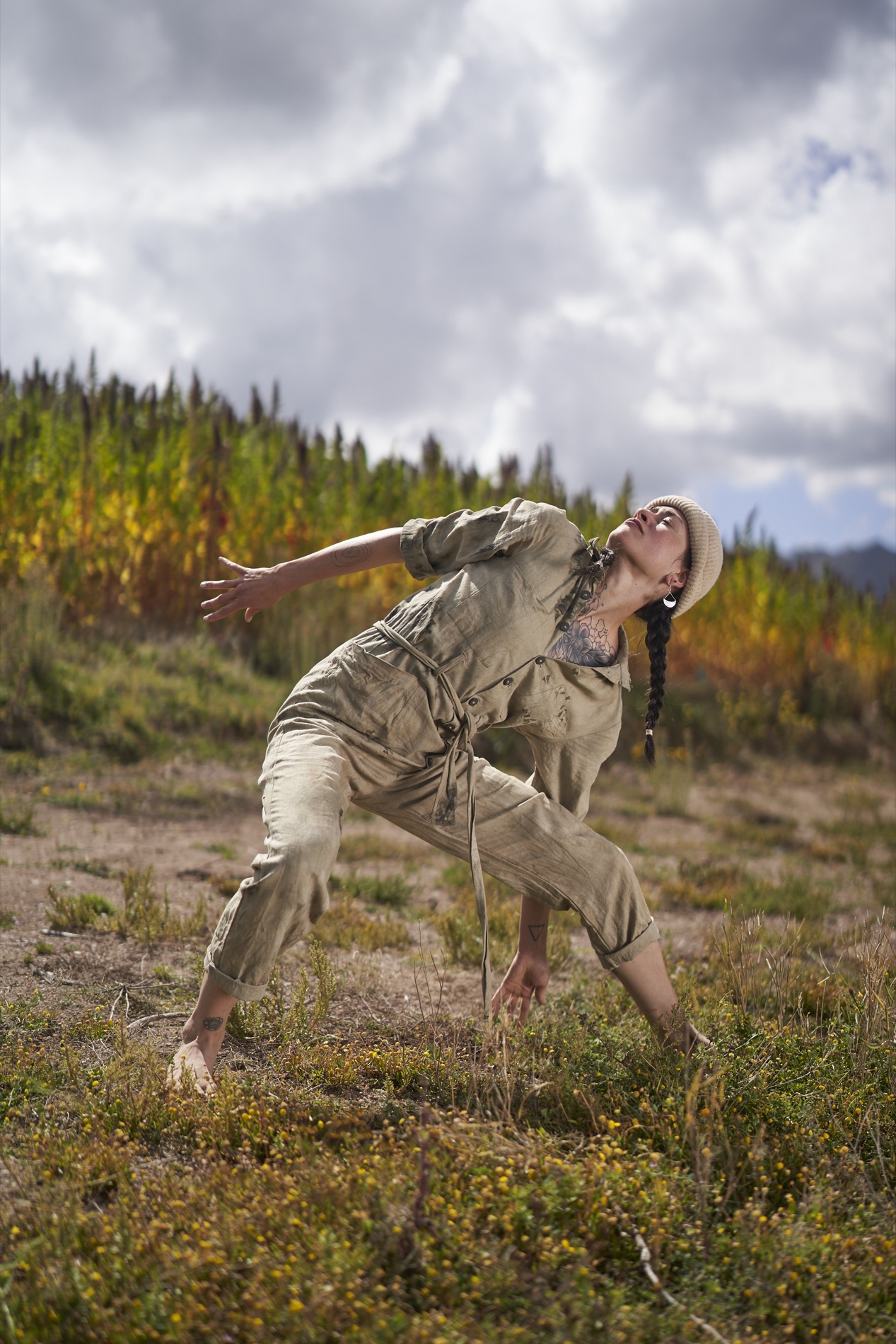 Dynamic outdoor portrait of a barefoot woman in a tan coverall arching backward in a dance pose in a field before a tall quinoa crop. Portrait by Alexander Estrada.