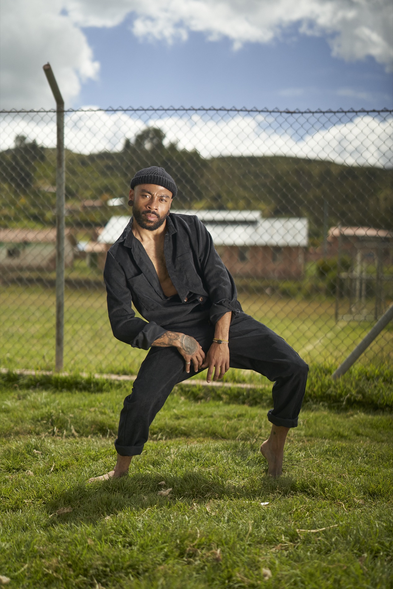 Environmental portrait of a bearded man in a black corduroy jacket and beanie crouching barefoot on grass in front of a chain-link fence and rural houses.