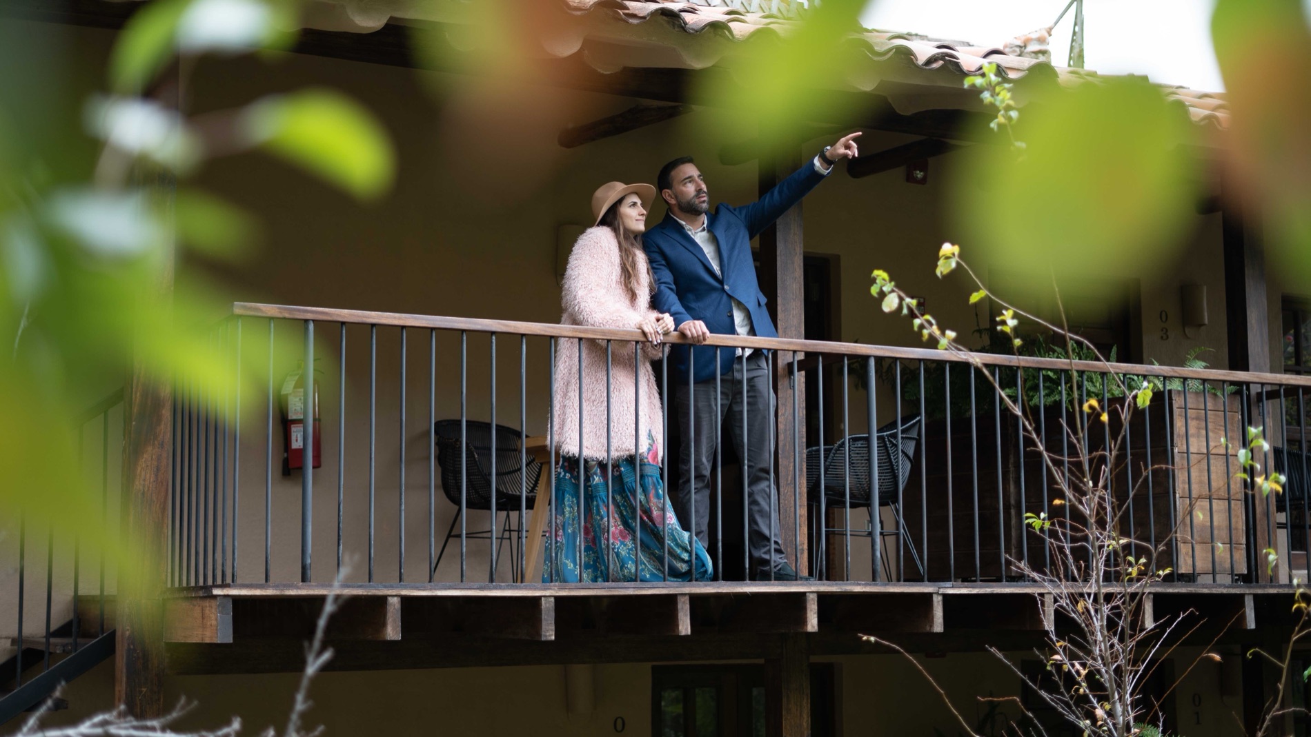 Documentary portrait of a couple standing on a wooden balcony of a tiled-roof lodge, the man pointing off into the distance, framed by green leaves.