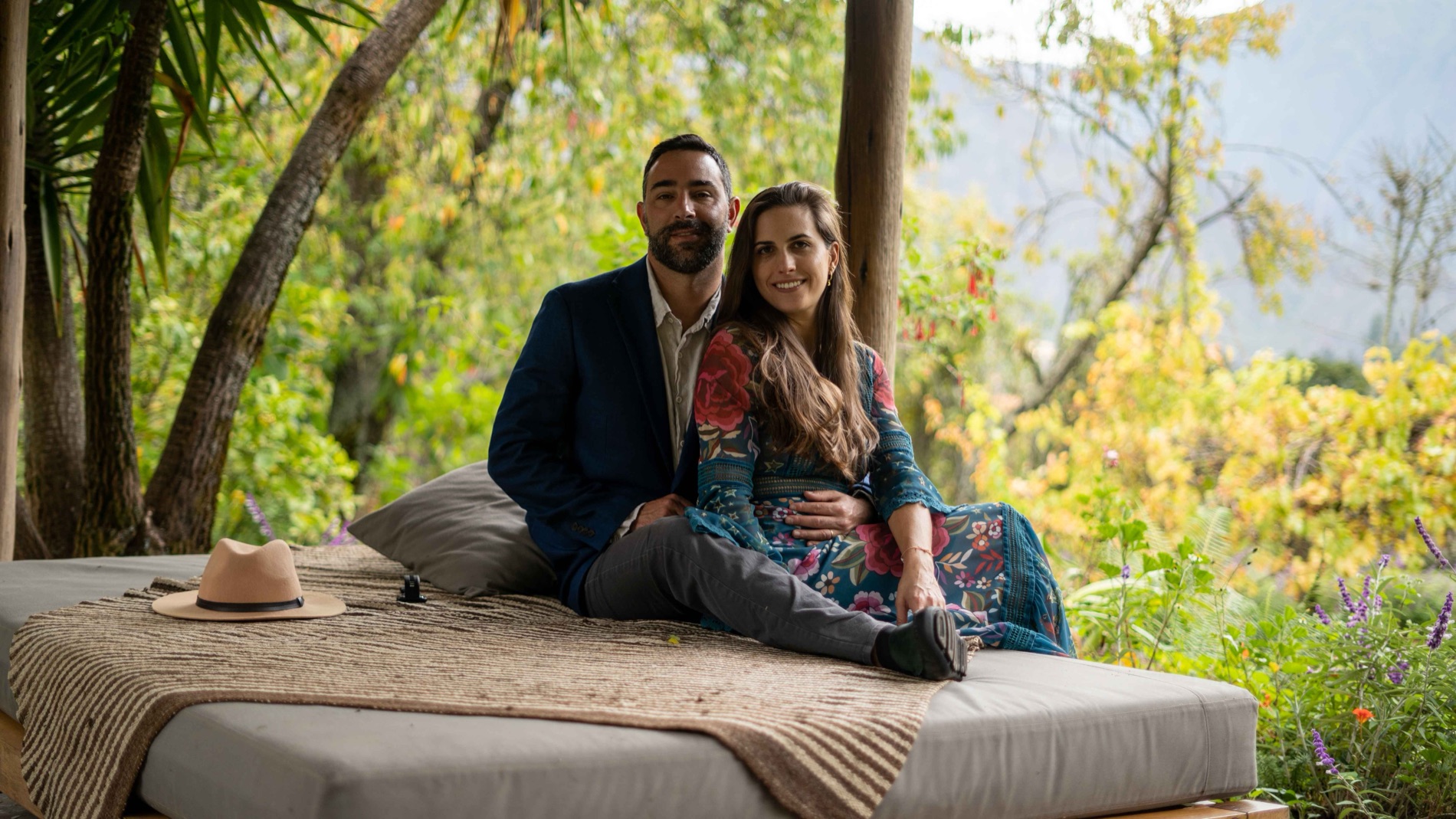 Outdoor portrait of a couple sitting together on a large daybed surrounded by lush green foliage and mountains, the man in a navy blazer and the woman in a floral dress. Portrait by Alexander Estrada.