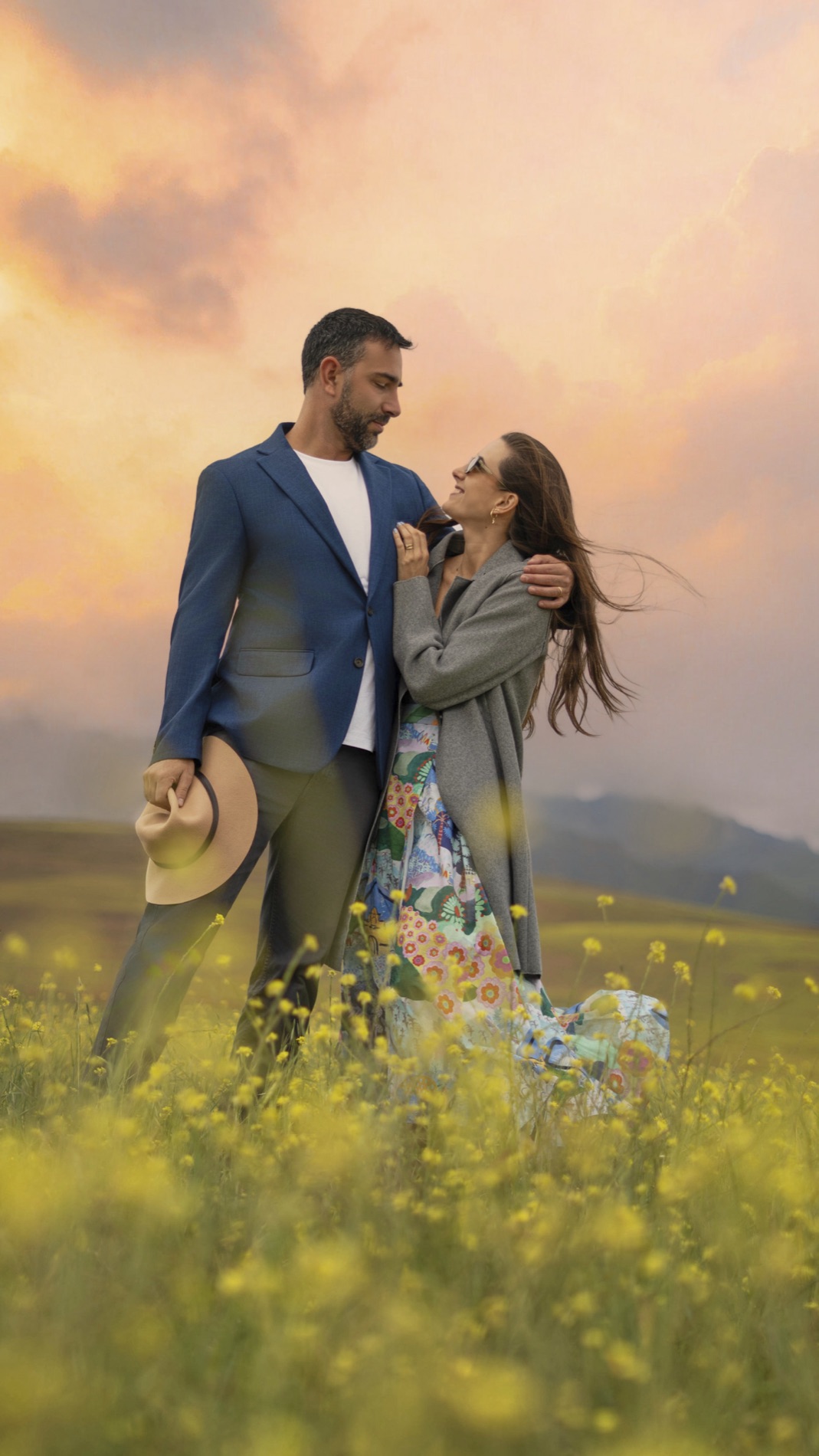 Editorial portrait of a couple embracing in a field of yellow flowers under a pink sunset sky, the man in a blue blazer and the woman in a colorful dress.