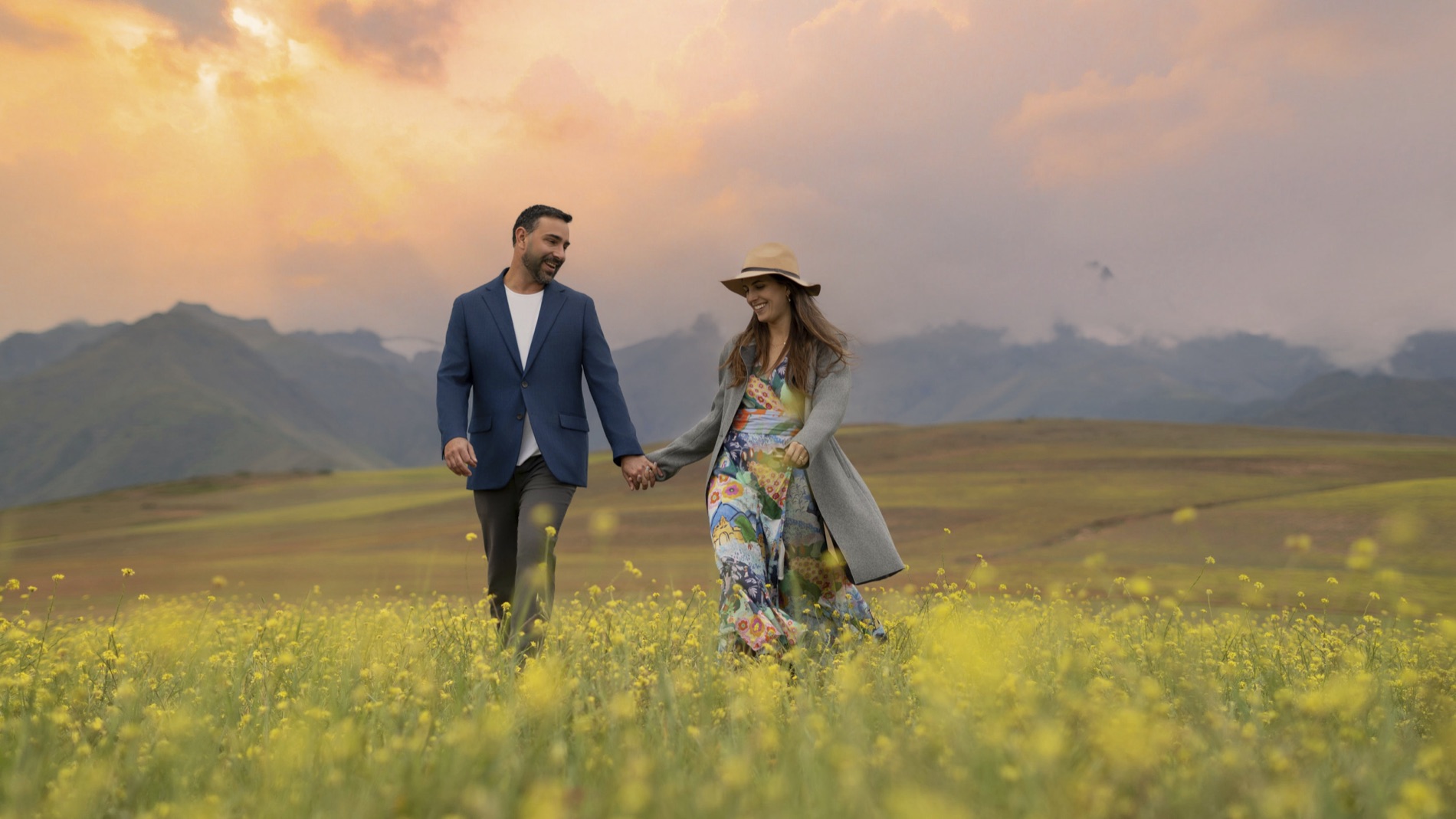 Outdoor portrait of a smiling couple holding hands as they walk through a blooming yellow field with mountains behind them at sunset. Portrait by Alexander Estrada.