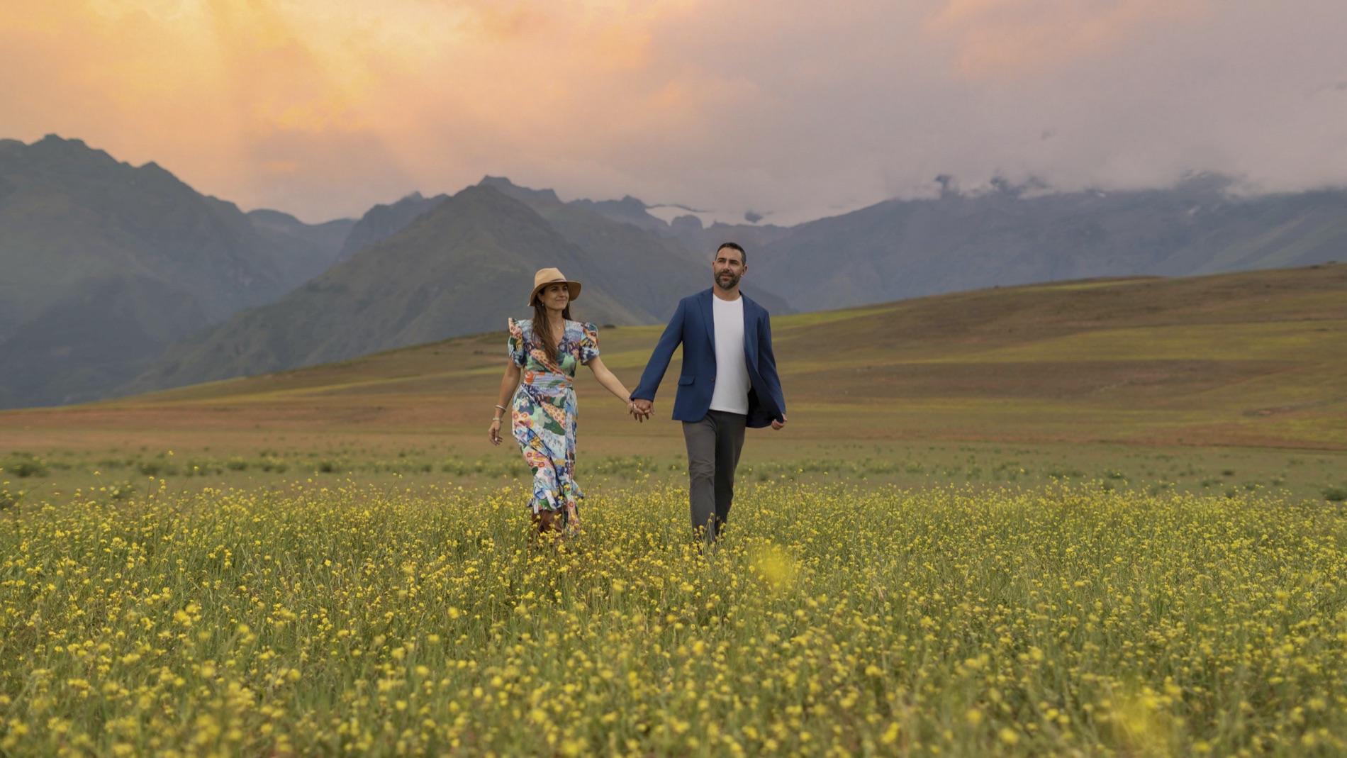 Wide environmental portrait of a couple holding hands walking through a yellow wildflower field below snow-capped mountains at sunset.